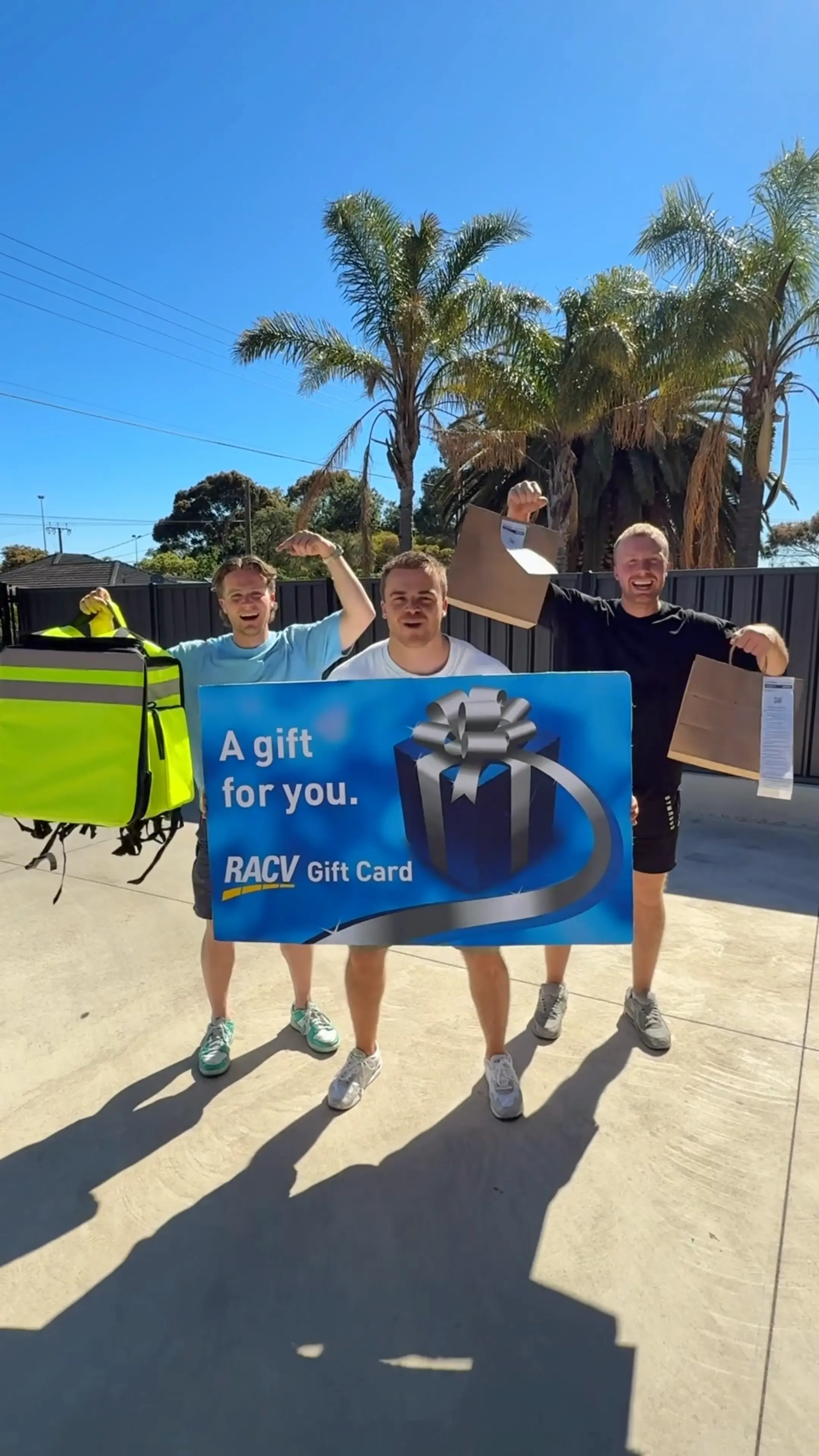 Three men outdoors smiling, holding a large blue RACV gift card and shopping bags under clear blue sky with palm trees in background.