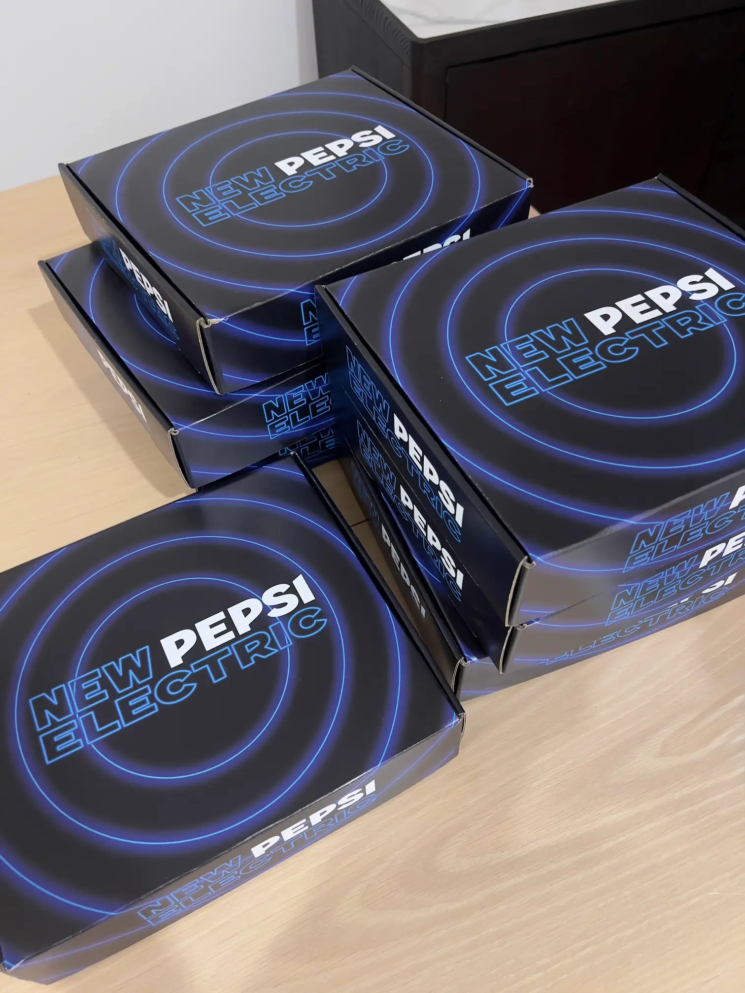 Stack of black boxes with blue circular designs labeled 'New Pepsi Electric' on a wooden surface.