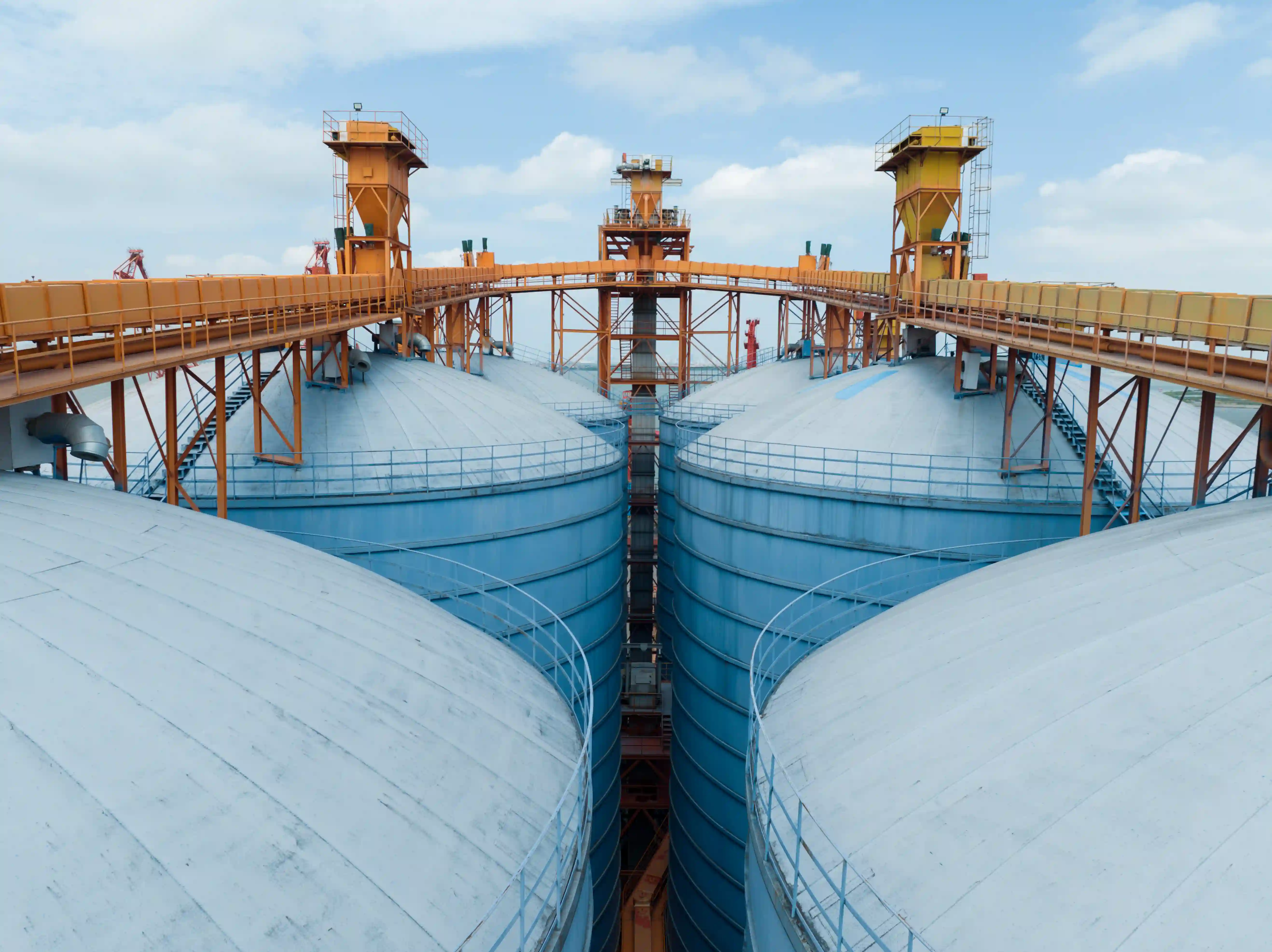 Luftaufnahme großer blauer Industriesilos mit orangenförmigen Brücken und Förderbändern unter bewölktem Himmel.