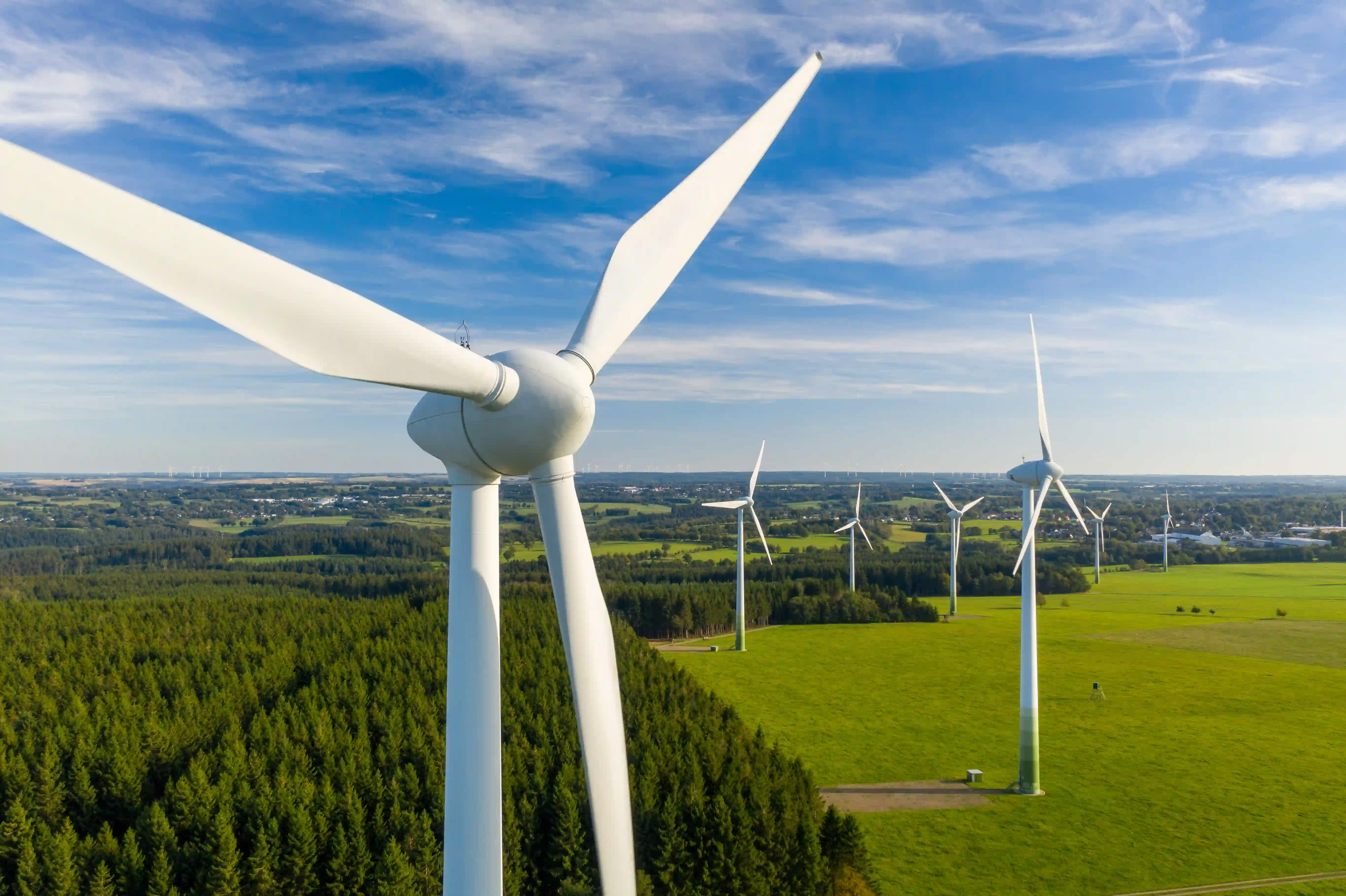 Windkraftanlagen auf grüner Wiese neben einem Nadelwald unter blauem Himmel mit Wolken.