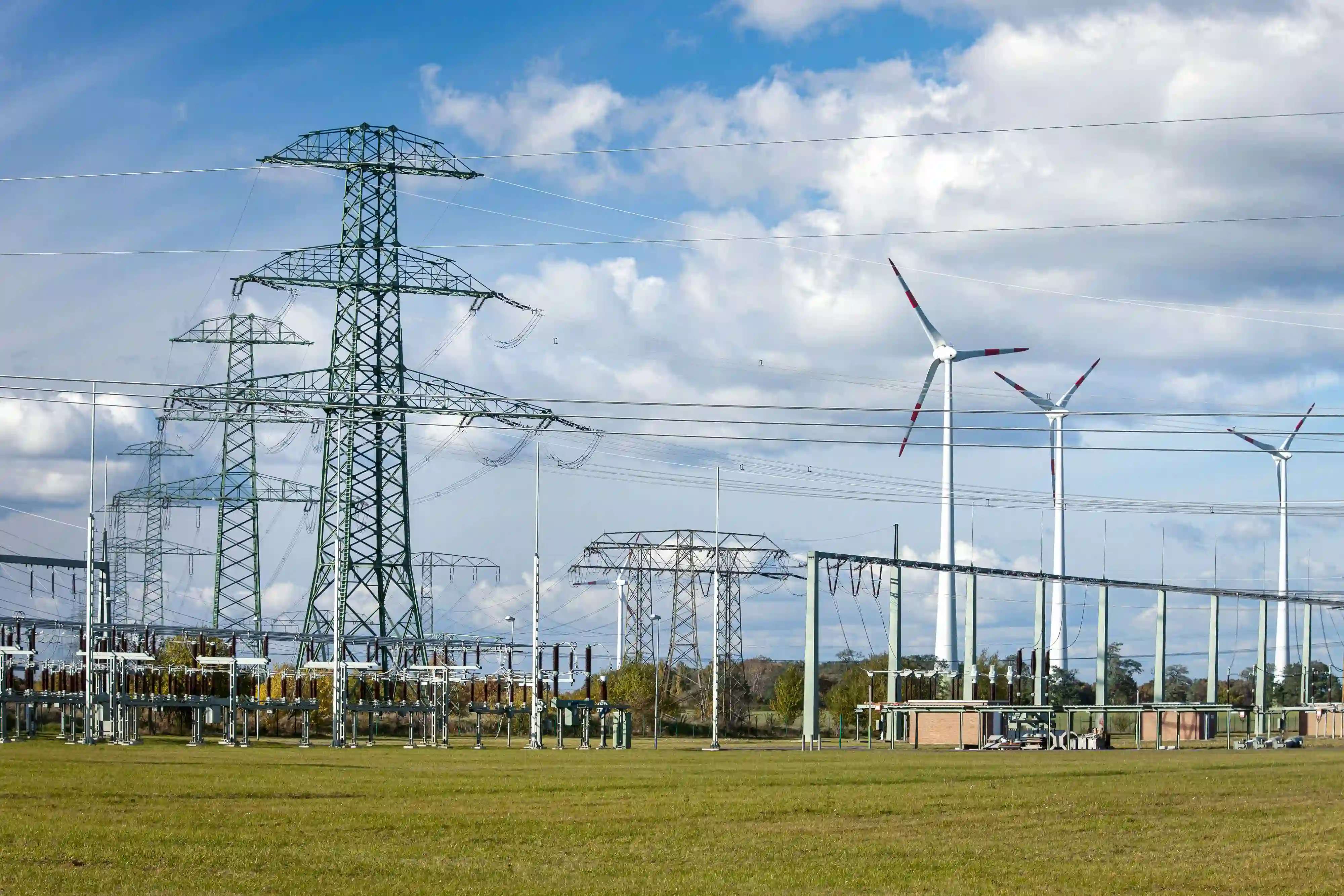 Feld mit Strommasten, Umspannwerk und Windkraftanlagen unter bewölktem Himmel.