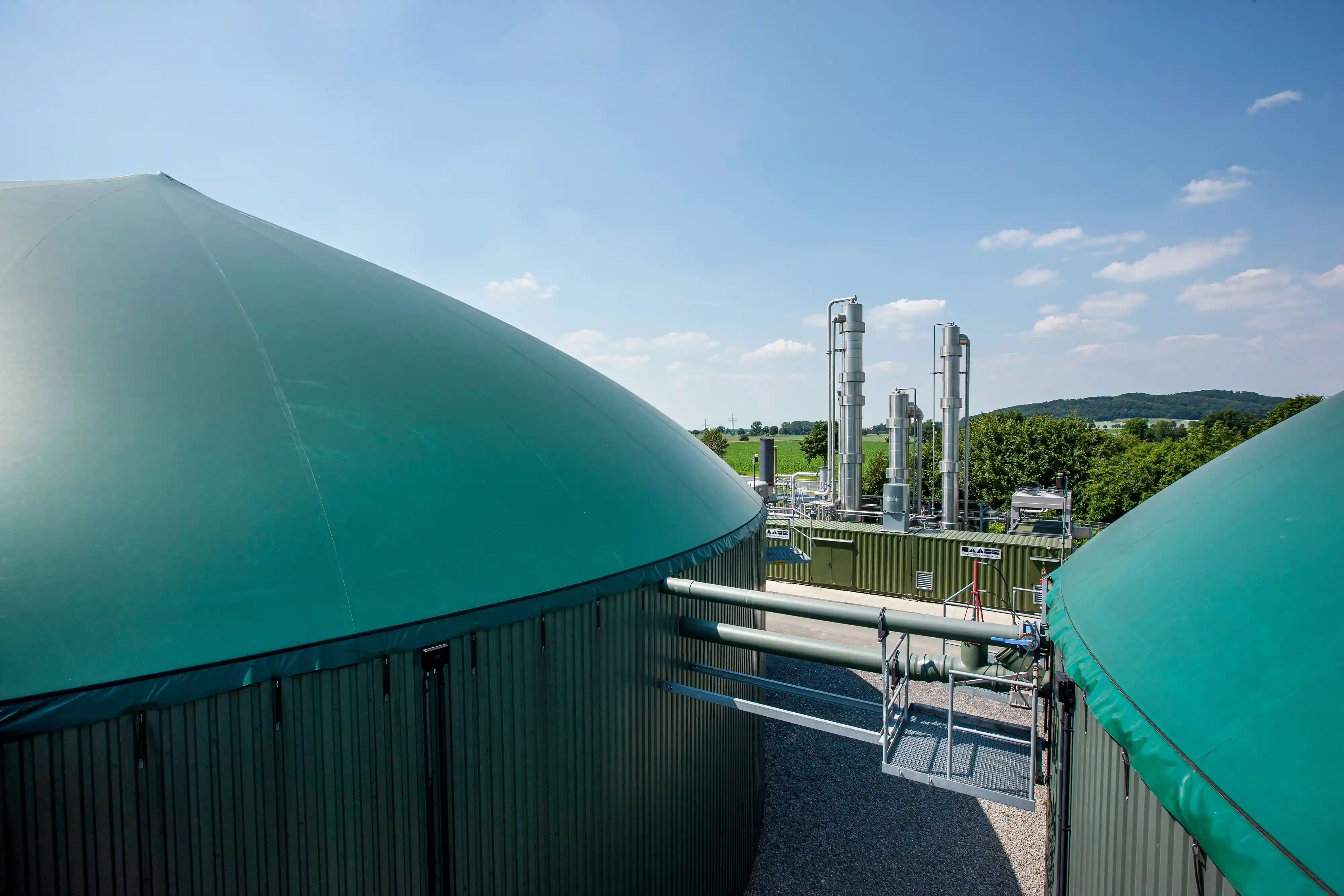 Zwei große grüne Biogastanks mit Metallrohren und technischem Equipment vor blauem Himmel und grüner Landschaft.