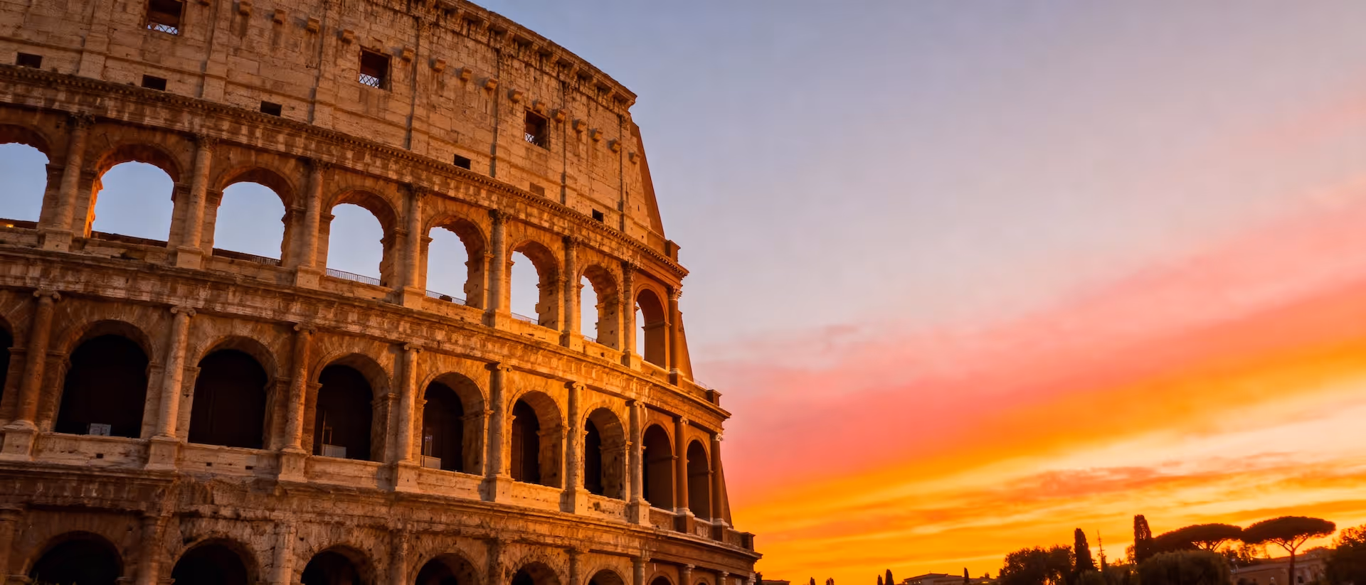 Partial side view of the ancient Roman Colosseum illuminated by a colorful orange and pink sunset sky.