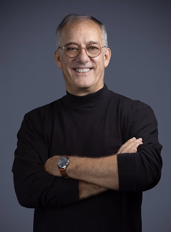 Smiling middle-aged man with glasses wearing a black turtleneck and watch, standing with arms crossed against a gray background.