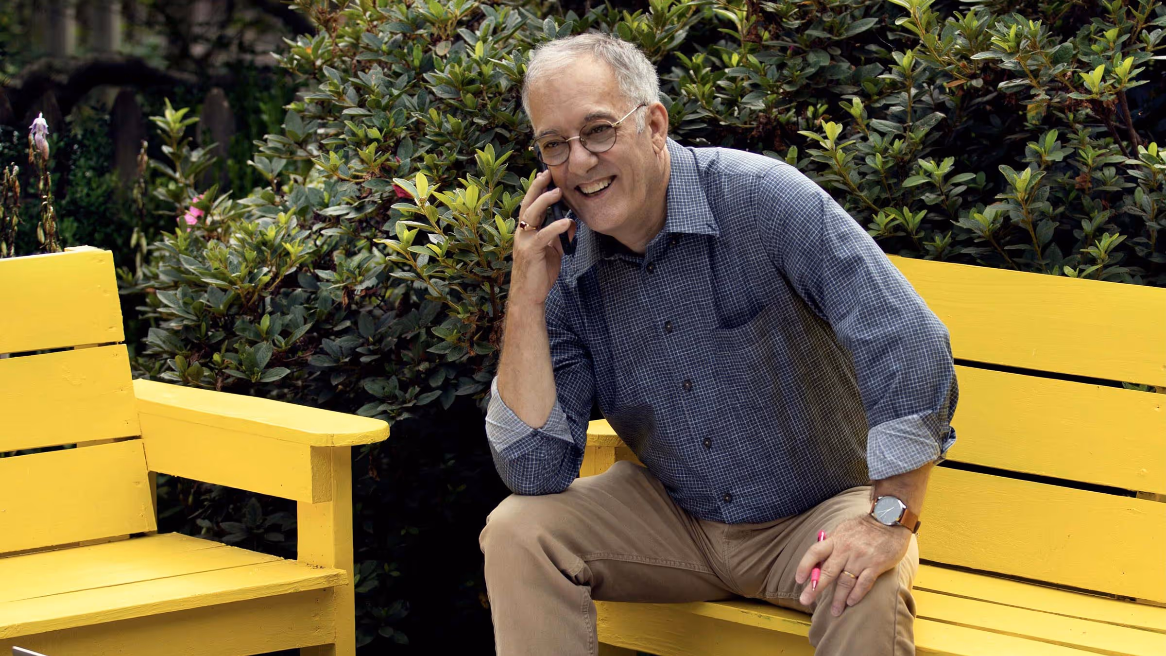 Smiling man wearing glasses and a blue checkered shirt, sitting on a yellow bench talking on a mobile phone surrounded by green bushes.