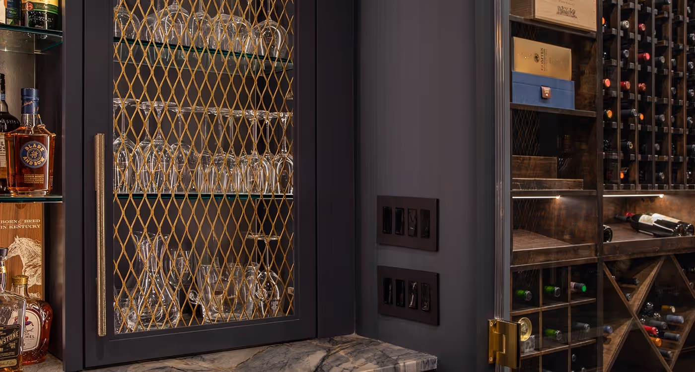 Close-up of a dark wooden liquor cabinet with glassware behind a decorative mesh door and wine bottles stored in racks beside it.