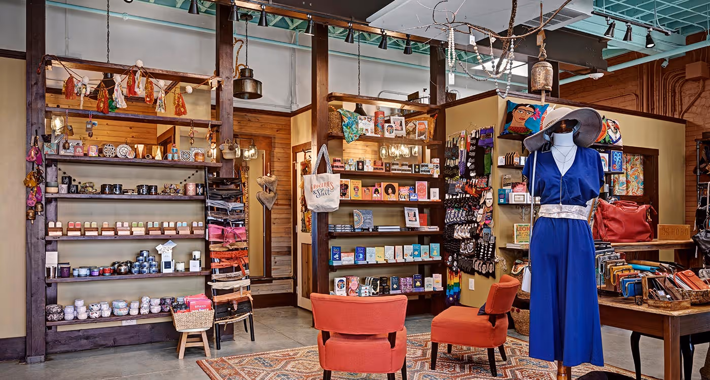Boutique interior with wooden shelves displaying skincare products, books, and accessories, orange chairs, and a mannequin dressed in a blue outfit and large hat.