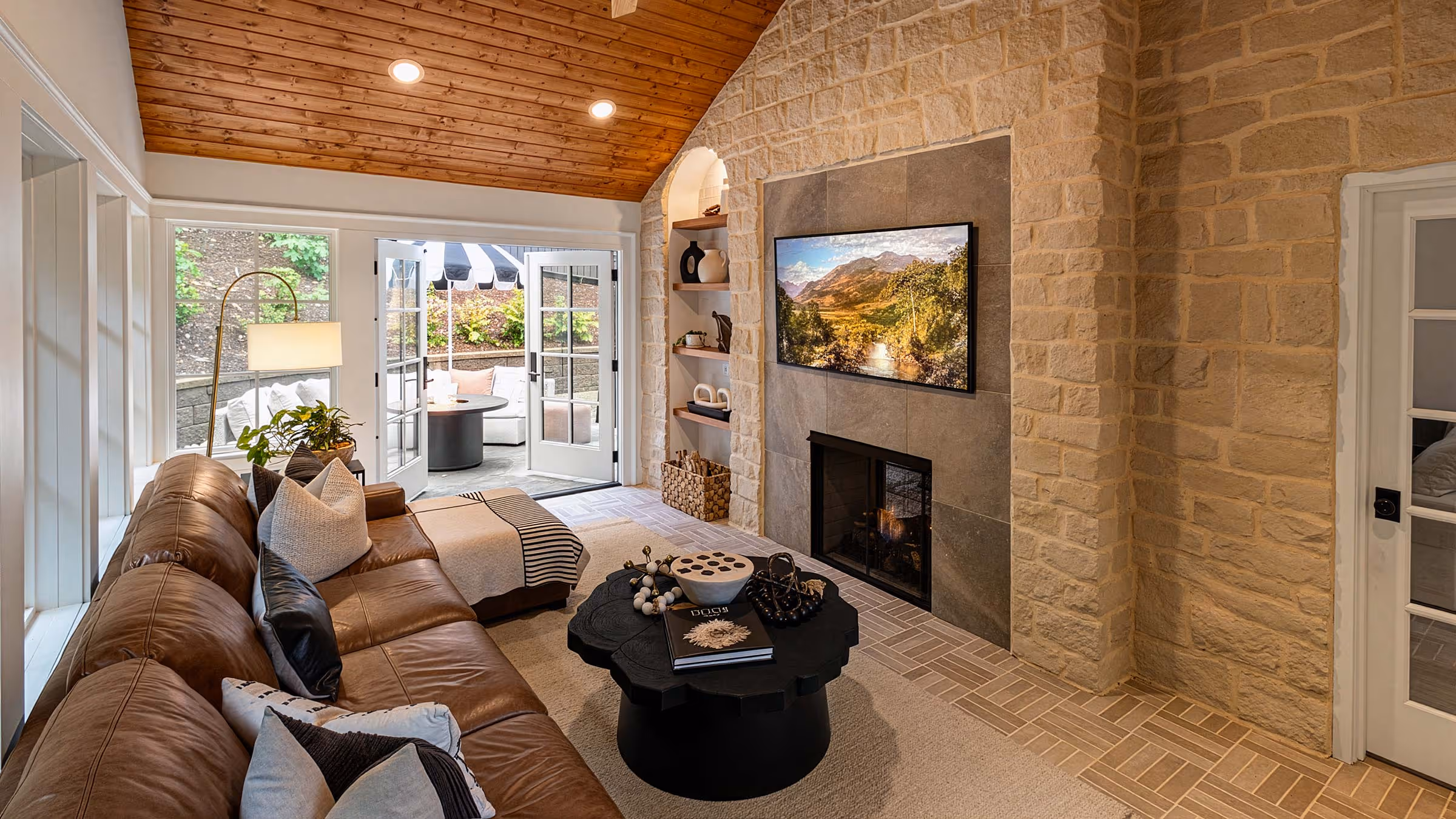 Cozy living room with a brown leather sectional, a modern fireplace set in a stone wall, a black round coffee table, and open glass doors leading to an outdoor seating area.