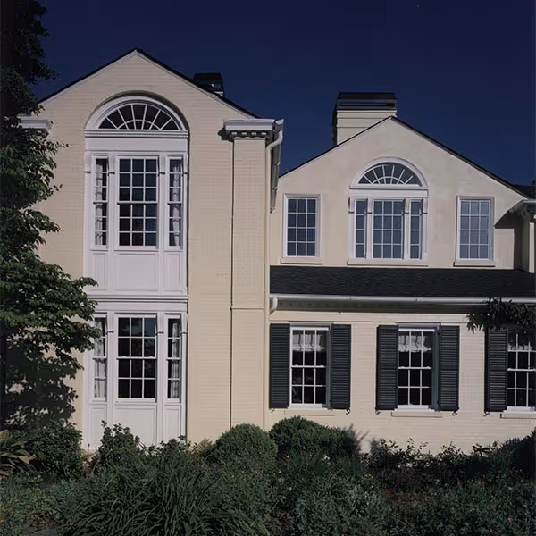 Two-story beige house with multiple white-framed windows and dark shutters against a night sky.