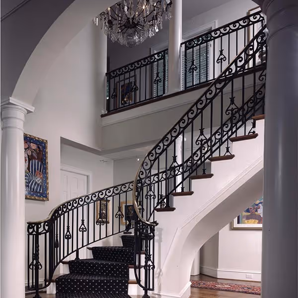 Interior view of a curved staircase with ornate black wrought iron railing and dark carpet with white dots, under white arches and columns in a bright room.