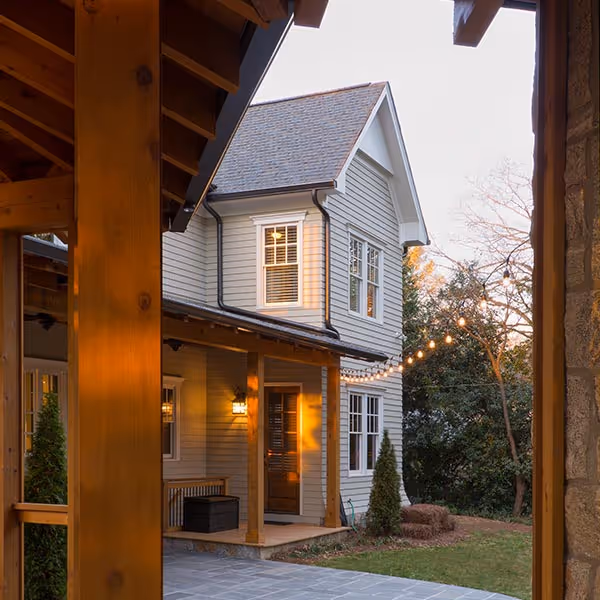 Two-story gray house with white trim seen through a wooden porch frame at sunset, with string lights and a small garden.