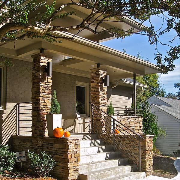Front porch of a house with stone pillars, concrete steps, two pumpkin decorations, and foliage around.