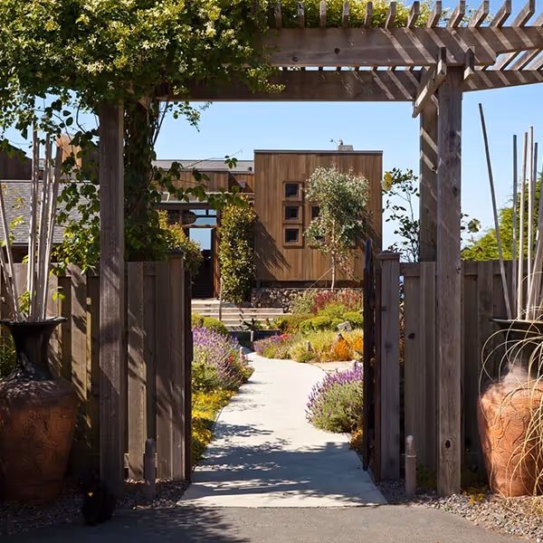 Garden pathway leading through a wooden gate with a pergola, surrounded by green plants and large decorative pots, toward a modern wooden house.