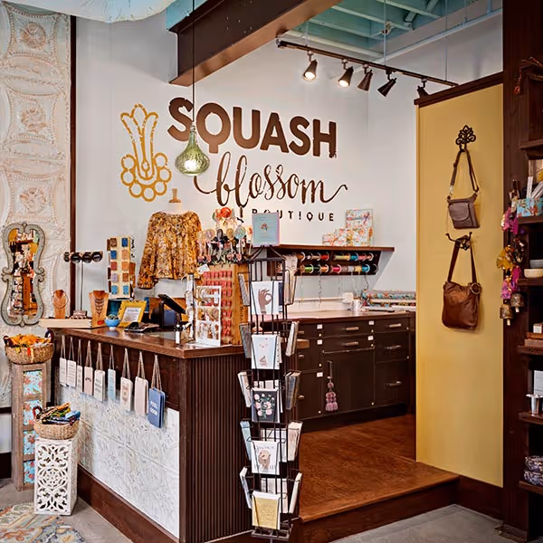 Cozy boutique interior with wicker chairs, floral arrangements, gold shelving displaying candles and beauty products, framed fashion artwork, and a statue by a table with white tablecloth.