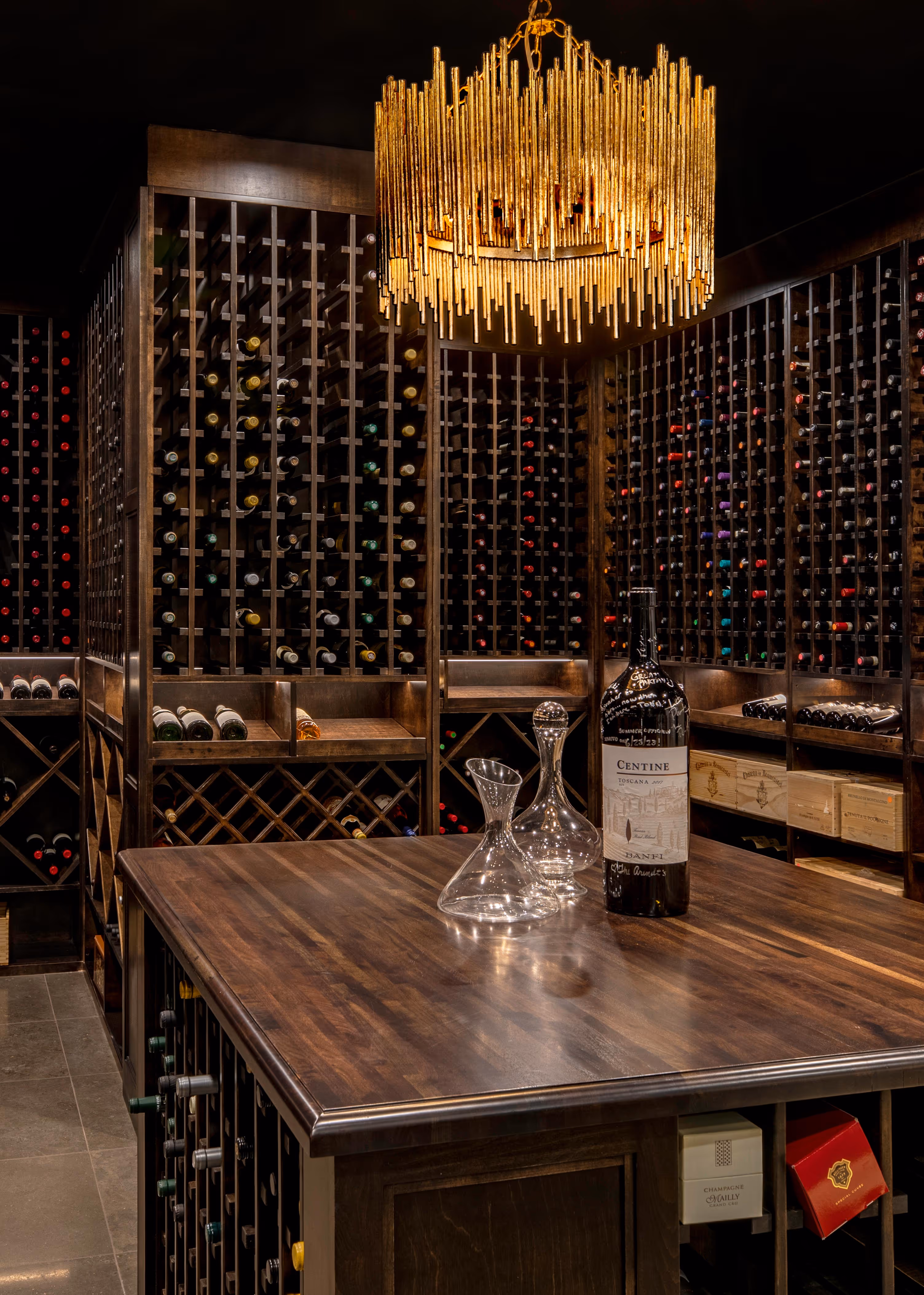 Luxurious wine cellar with dark wood racks filled with wine bottles and a wooden island table topped with a wine bottle and glass decanters under a modern gold chandelier.