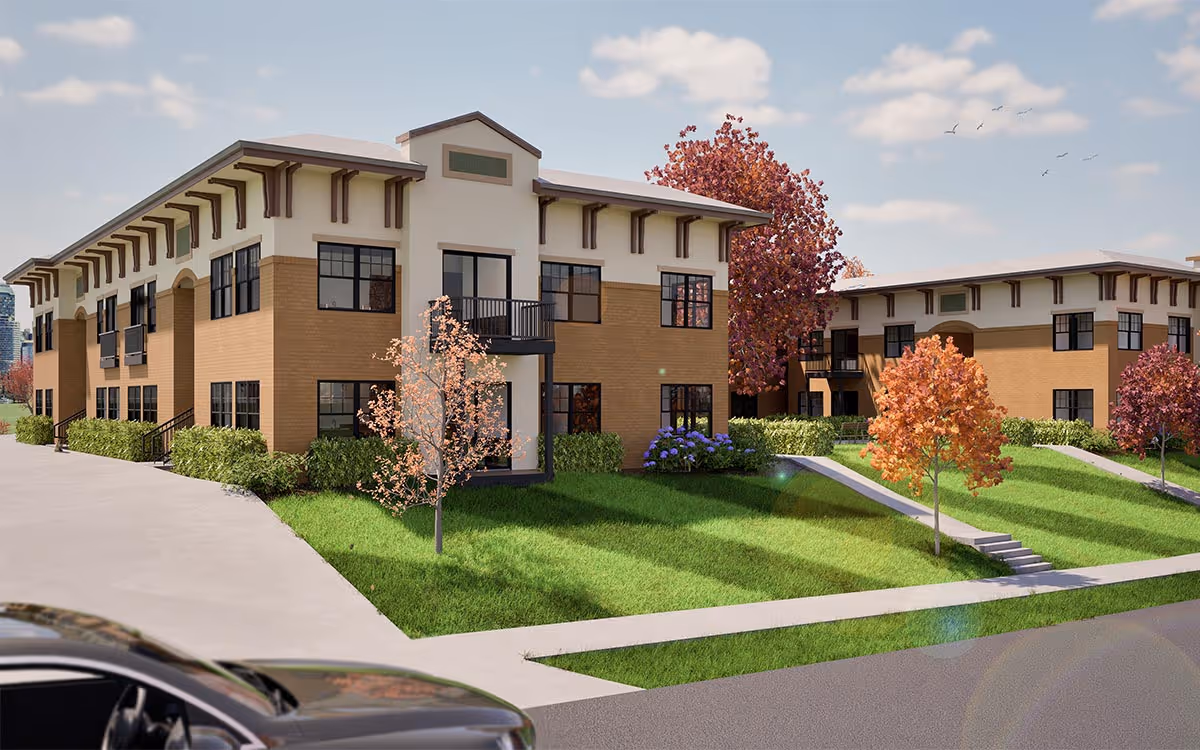 Two-story beige and brick apartment buildings surrounded by green lawns and autumn trees with orange and purple leaves under a partly cloudy sky.