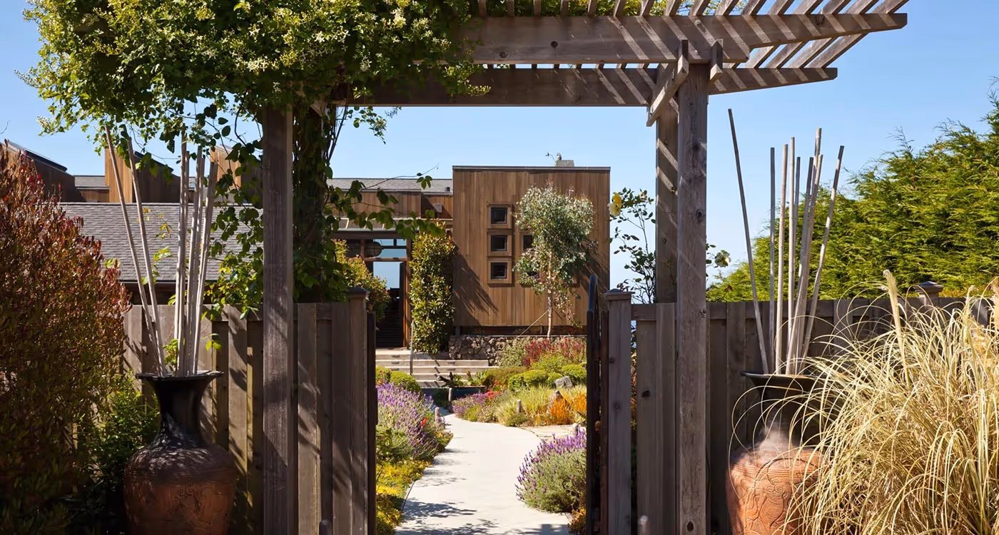 Wooden pergola entrance leading to a garden pathway surrounded by plants and shrubs, with a modern wooden house in the background under a clear blue sky.