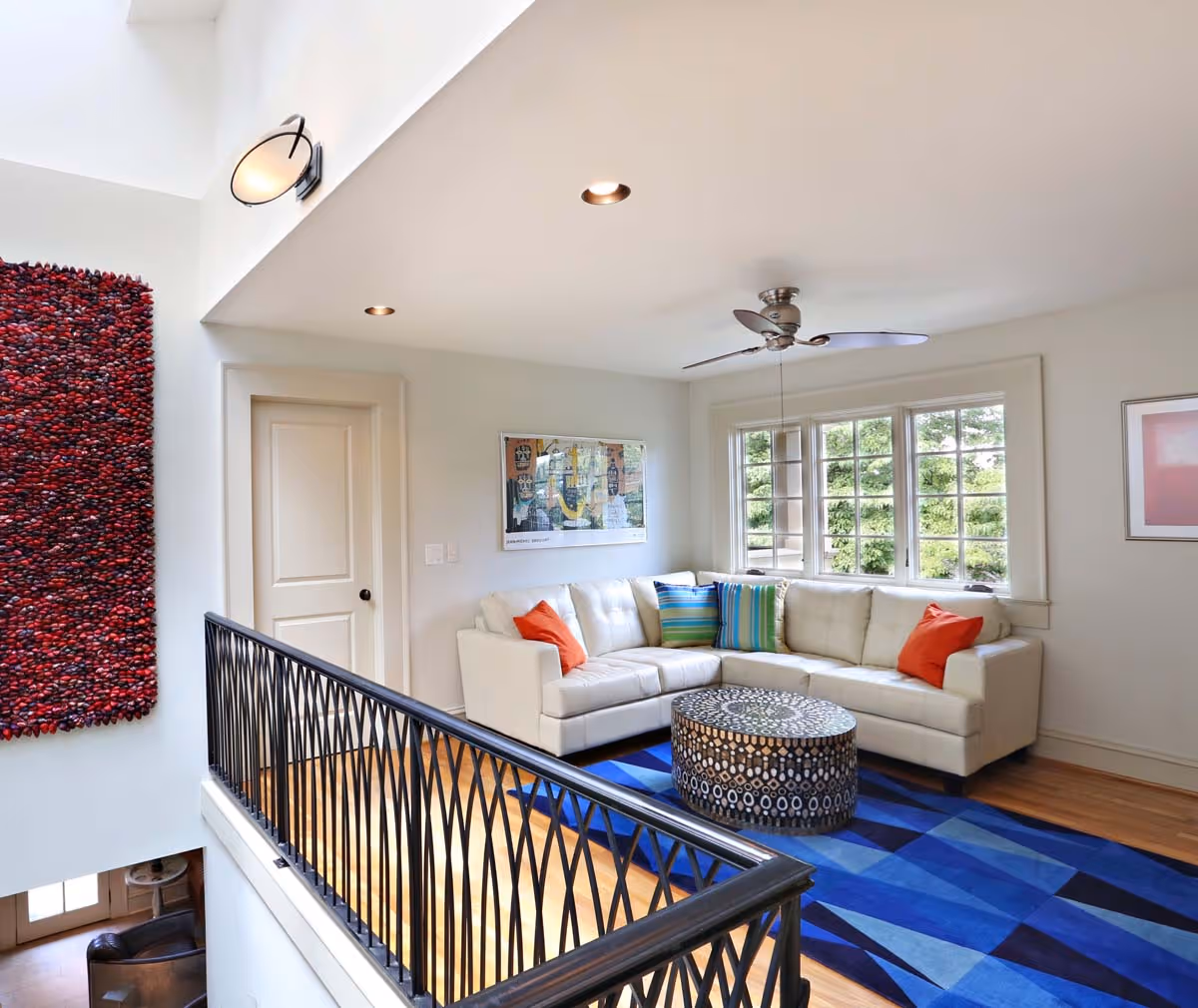 Bright living room with large windows, a grand piano, gray armchair, beige sofa with colorful pillows, and a coffee table with flowers and books.