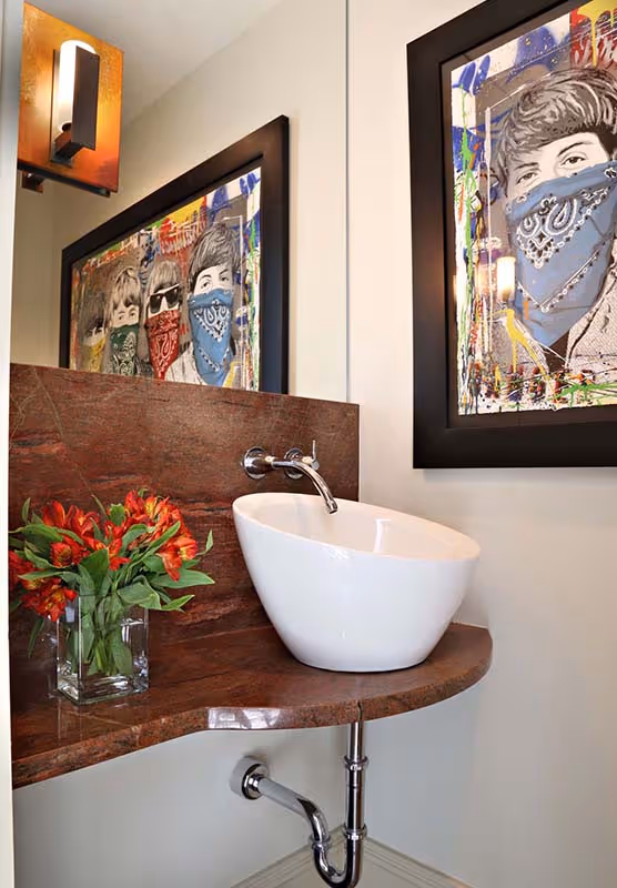 Modern bathroom sink with a white oval vessel basin on a curved brown marble countertop, a chrome wall-mounted faucet, red flowers in a clear vase, and colorful artwork of masked faces on the wall.