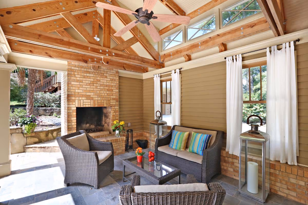 Cozy outdoor patio with wicker furniture, striped cushions, brick fireplace, and a ceiling fan under exposed wooden beams.
