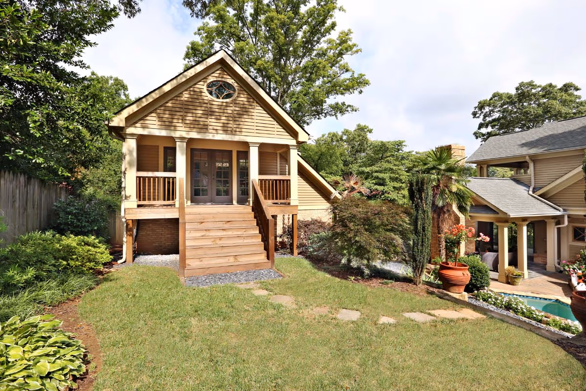 A small wooden cottage with stairs leading to a porch, surrounded by green grass, shrubs, and trees, next to a larger house with a swimming pool.