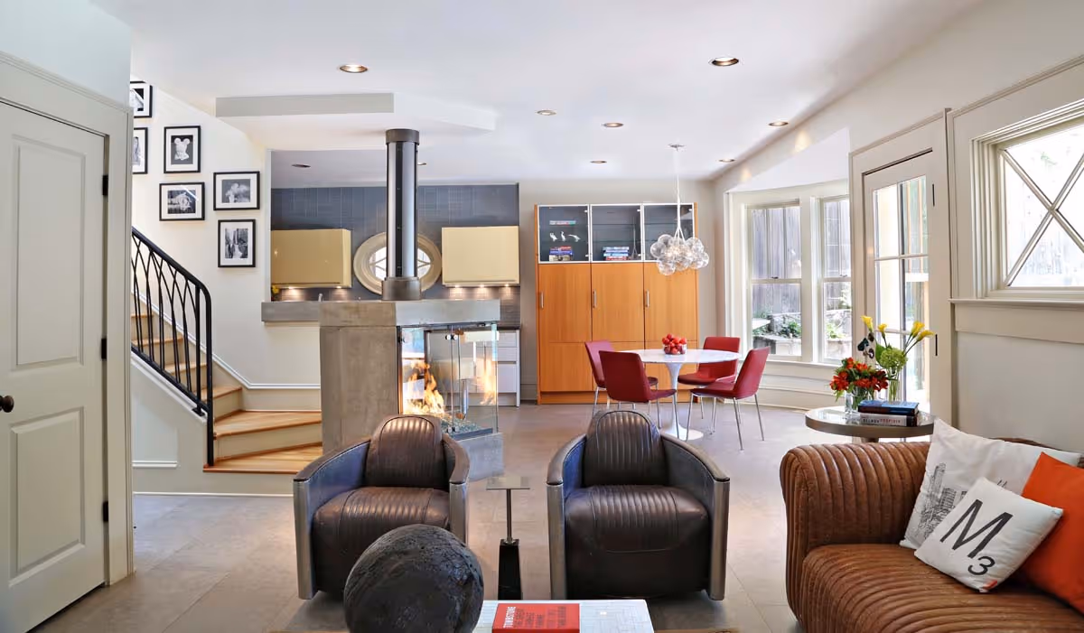 Bright loft area with leather bench, dark wood bookcase with glass doors, armchairs, indoor plants, and wooden floor.