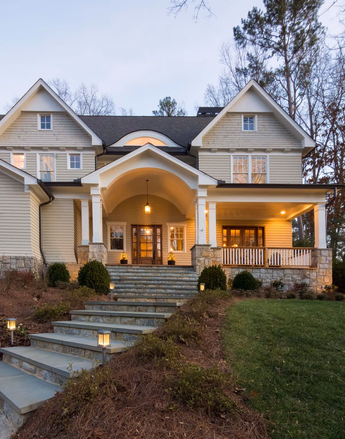 Bright living room with a tall potted plant beside a fireplace, two dark armchairs, a large painting above the fireplace, and an adjacent seating area with a white armchair and brown sofa.