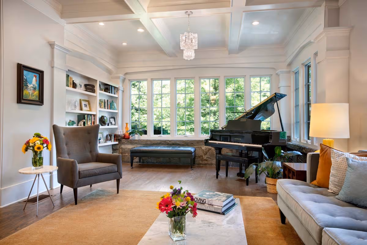 Bright living room with large windows, a black grand piano, grey armchair, beige sofa, and floral arrangements on tables.