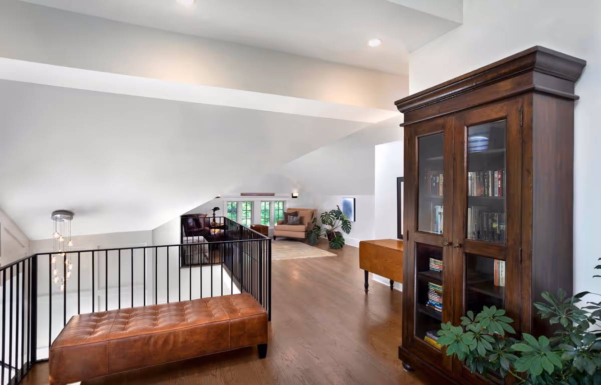 Bright loft with brown leather bench, dark wooden bookcase filled with books, beige armchair, plants, and hardwood floors.