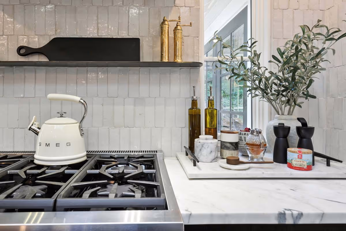 Modern three-story townhouses with white siding and large windows under a blue sky, surrounded by trees and with people walking nearby.