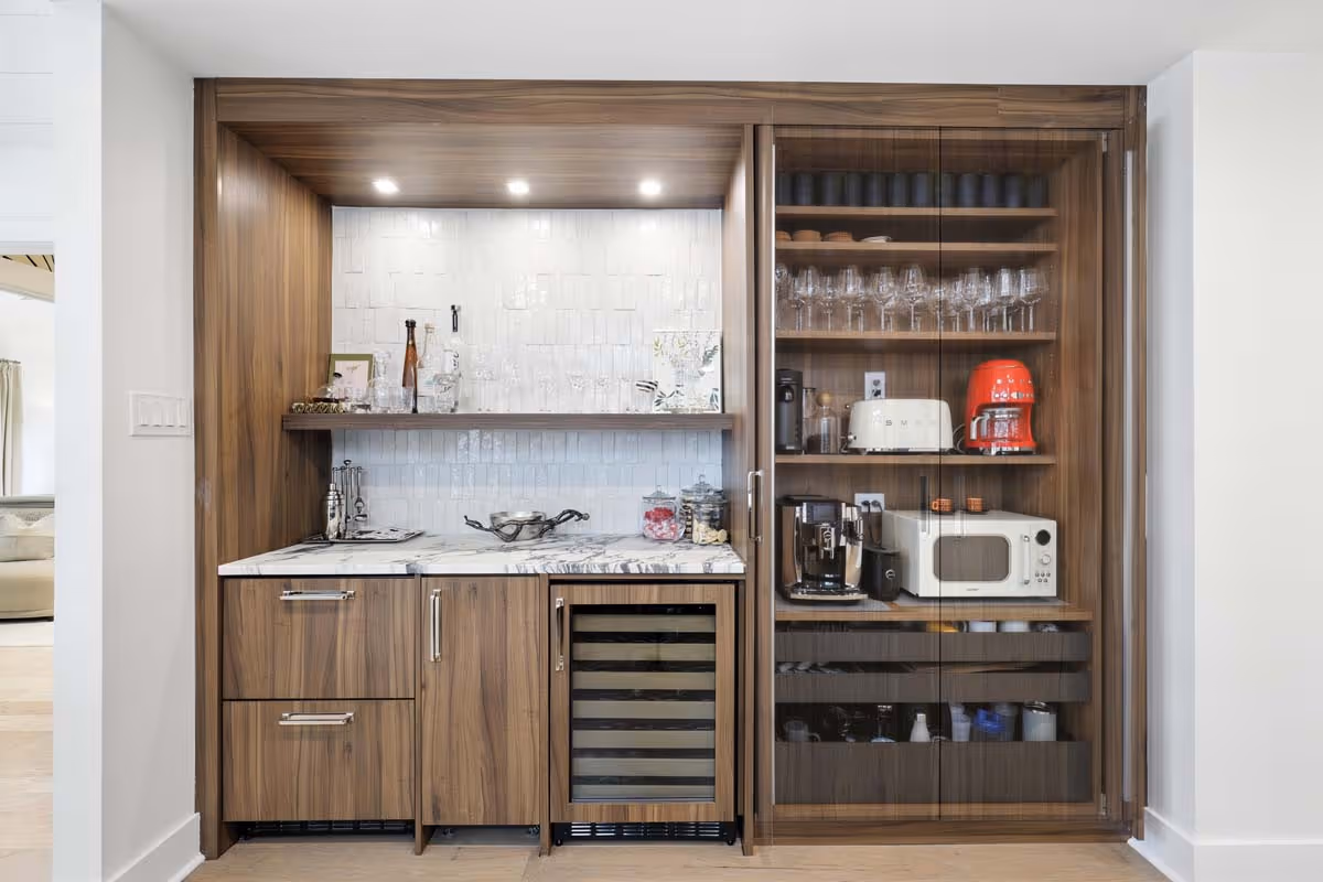 Modern wooden kitchen bar setup with marble countertop, wine cooler, glassware, coffee machines, toaster, microwave, and shelves behind glass doors.