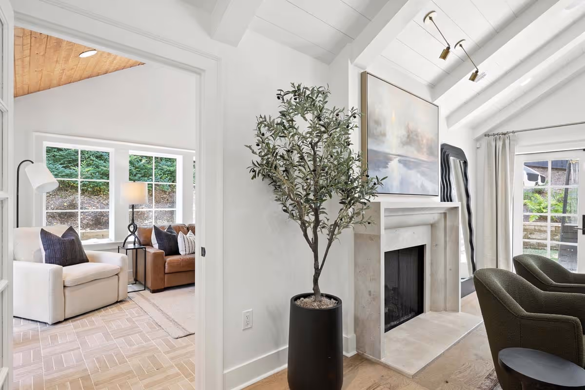 Modern living room with a tall potted plant beside a stone fireplace, armchairs, sofa, and large windows.