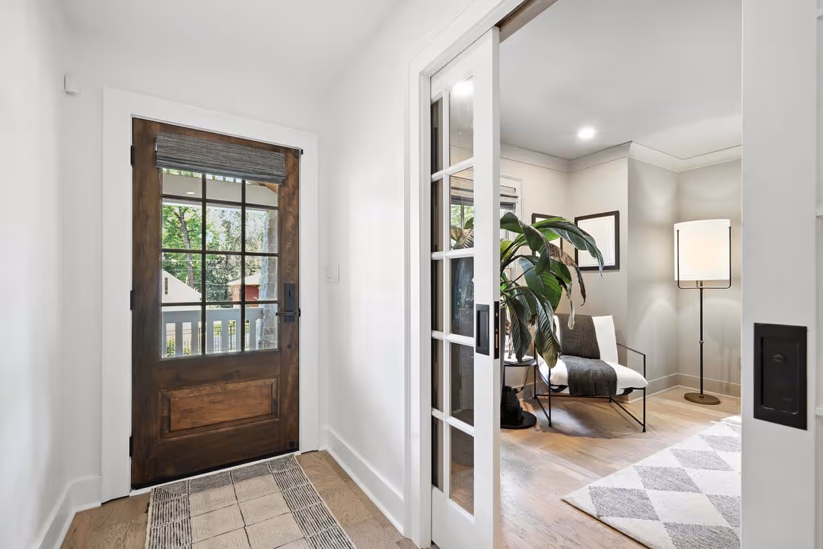 Cozy living room with a gray sectional sofa adorned with red and beige pillows, a glass coffee table with decorative items, framed artwork on the wall, potted plants near a large window, and a red vase of sunflowers on a side table.