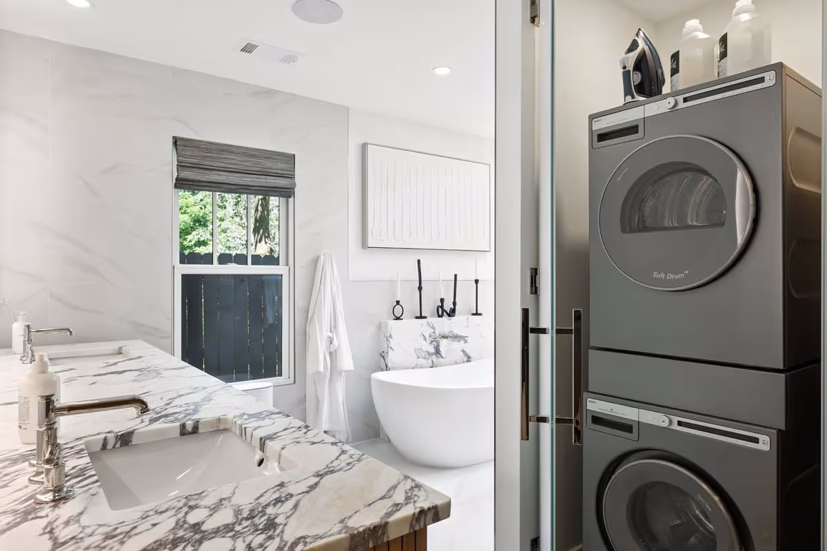 Modern bathroom with marble countertop sinks, freestanding bathtub, and stacked washer and dryer unit.