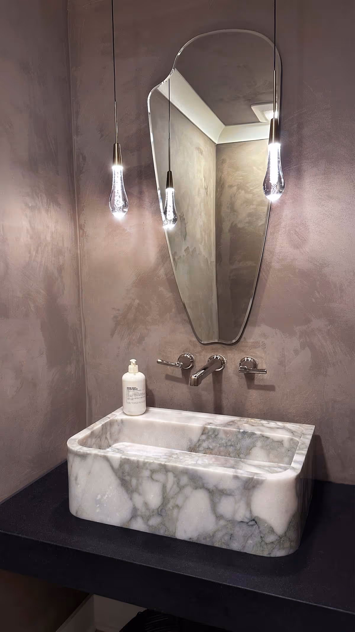 Modern bathroom sink made of white and gray marble with wall-mounted faucet, an irregularly shaped mirror, and two hanging pendant lights.