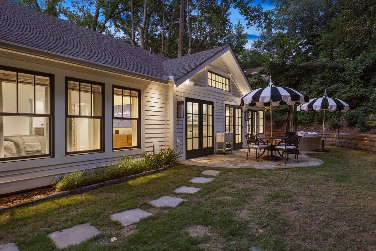 Backyard patio of a house with stone pavers, black and white striped umbrellas, outdoor seating, and surrounding trees at dusk.