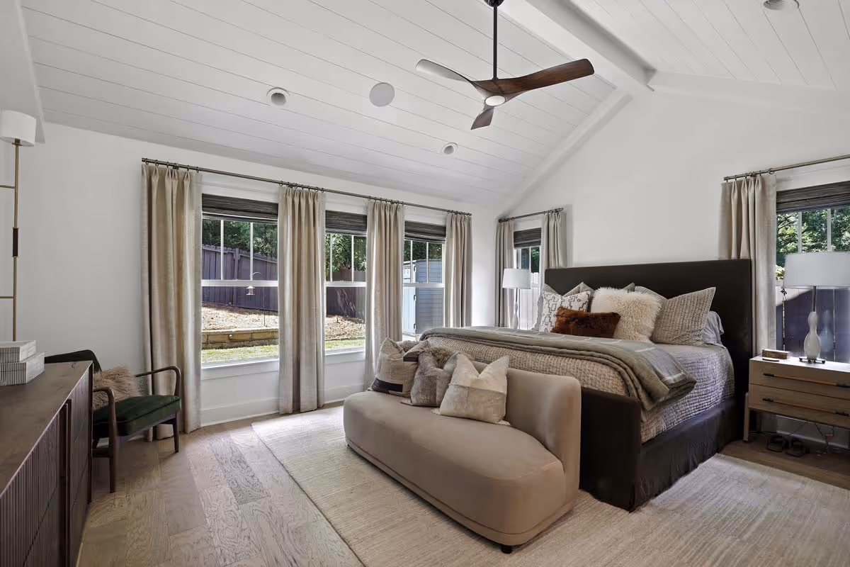 Bright modern bedroom with vaulted white ceiling, dark bed frame with multiple pillows, beige sofa, and large windows with beige curtains.