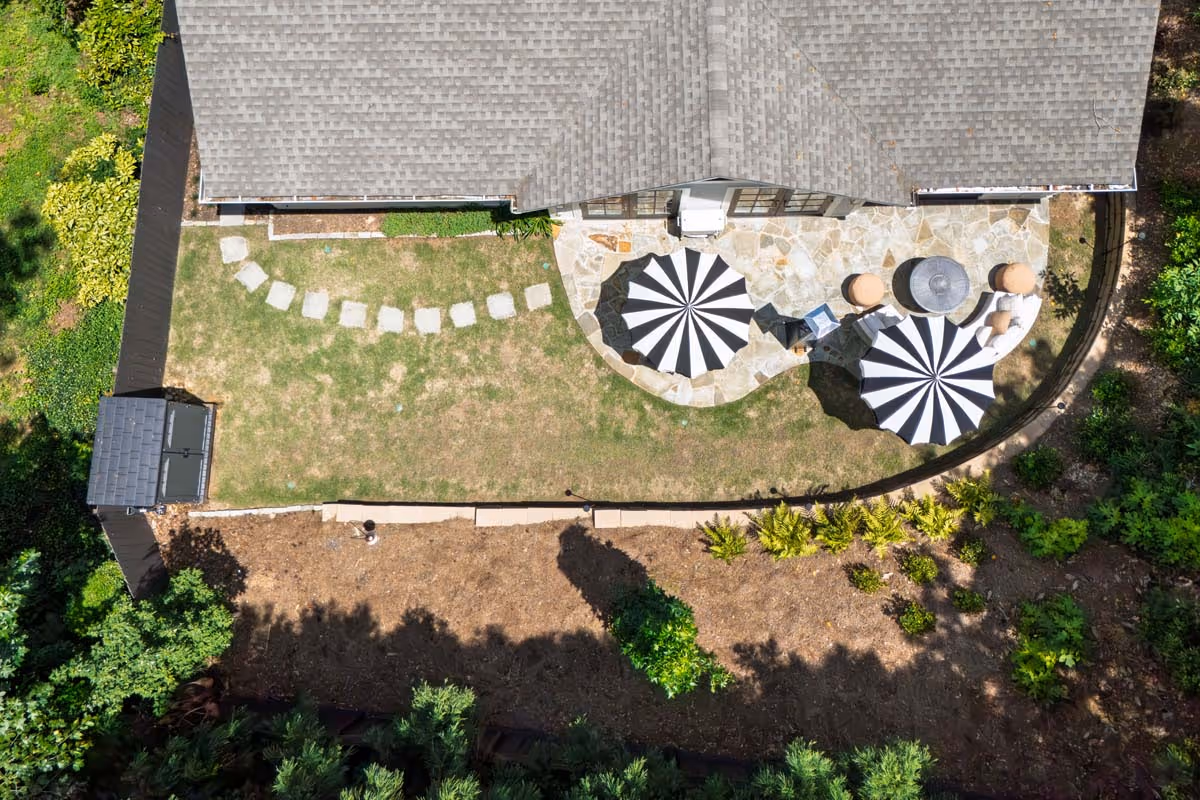 Aerial view of a backyard with a stone patio, two black-and-white striped umbrellas, outdoor seating, and a small garden area.