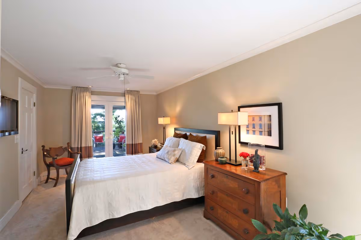 Bright living room with large windows, a grand piano, gray armchair, light gray sofa with cushions, flowers on a marble coffee table, bookshelf, and a table lamp.