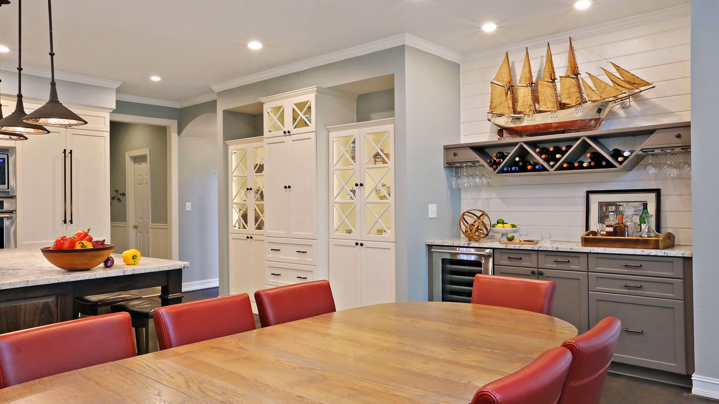 Modern dining area with wooden table, red chairs, kitchen island with vegetables, white cabinets, gray drawers, wine rack, and a large model sailboat.