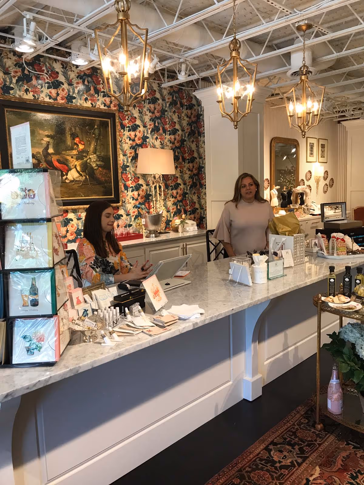 Cozy living room with gray sectional sofa, colorful pillows, glass coffee table with decorative items, indoor plants, and sunflower bouquet on a side table.