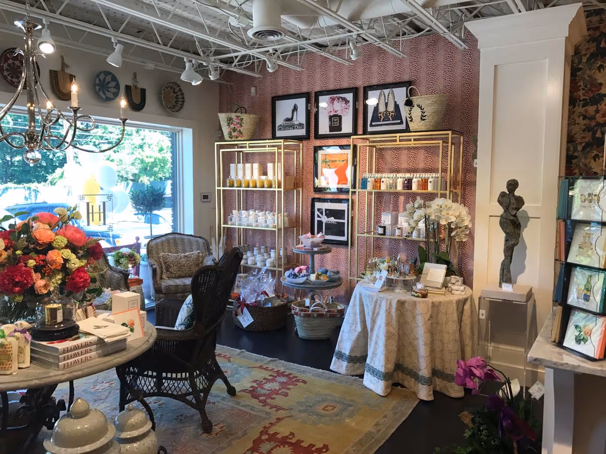 Cozy boutique interior with floral arrangements, wicker chairs, decorative shelves with beauty products, framed art on patterned wall, and a small table with orchids.