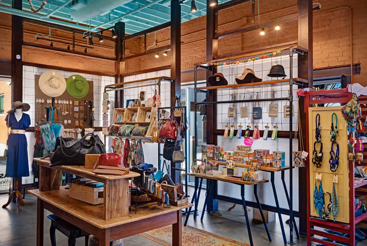 Modern living room with gray sectional sofa, red and white patterned pillows, glass coffee table with decorative items, plants near a large window, and a vase of sunflowers on a side table.