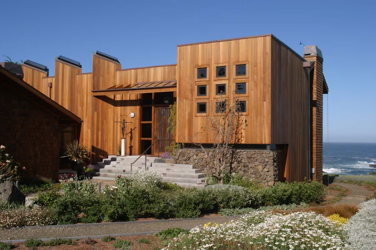 Two-story beige and brown apartment buildings with balconies, surrounded by green lawns and autumn-colored trees under a partly cloudy sky.