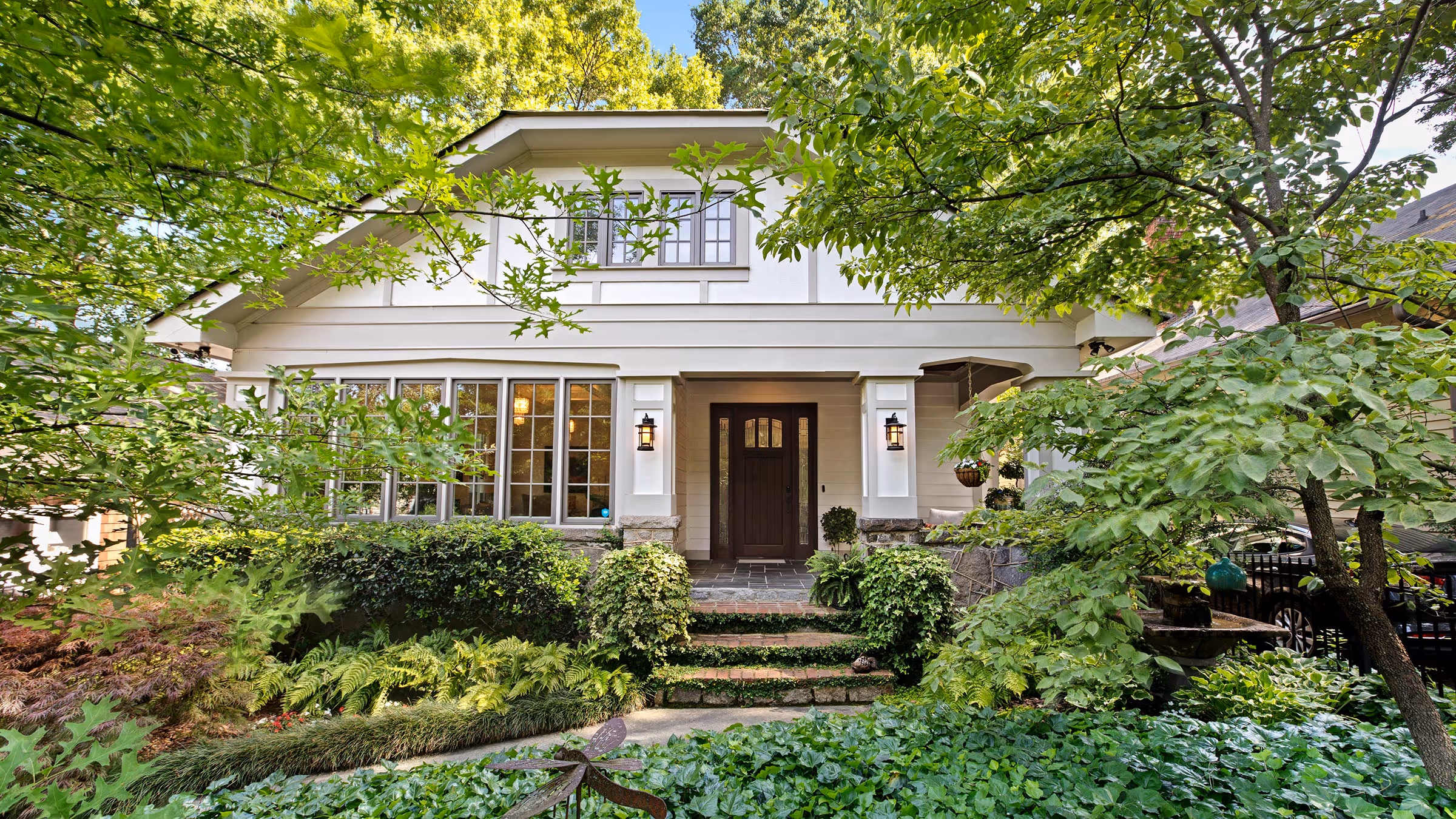 Sunlit porch with two mesh chairs, metal railing, and stone half-wall overlooking green trees and bushes.
