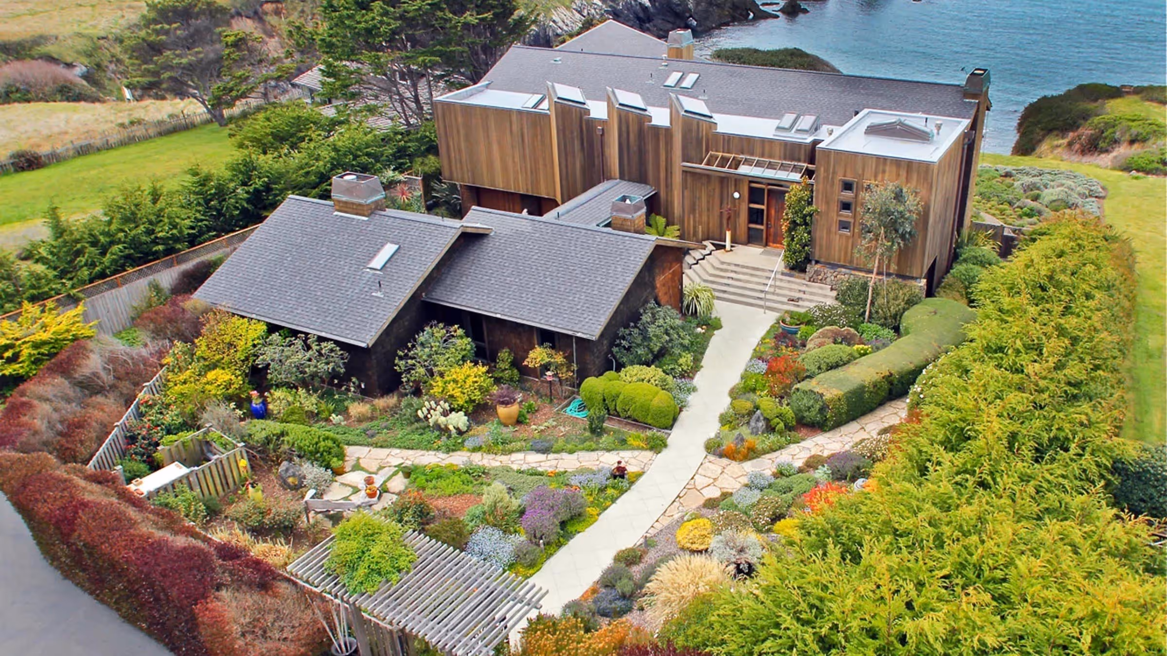 Aerial view of a modern wooden house with multiple gray roofs surrounded by a lush, colorful garden and overlooking a body of water.