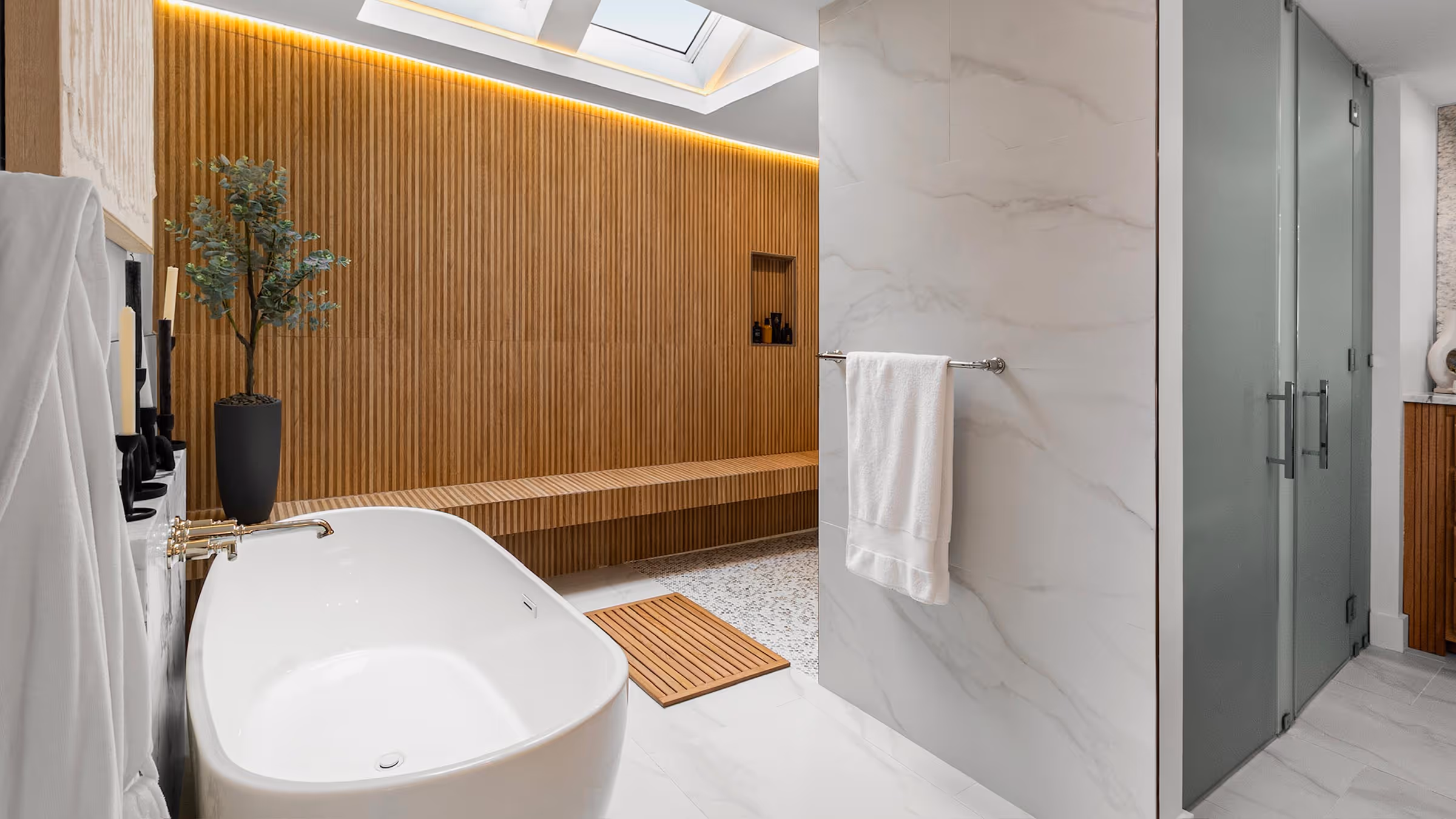 Modern bathroom with a freestanding white bathtub, wooden slatted wall and bench, a black potted plant, and frosted glass shower doors.