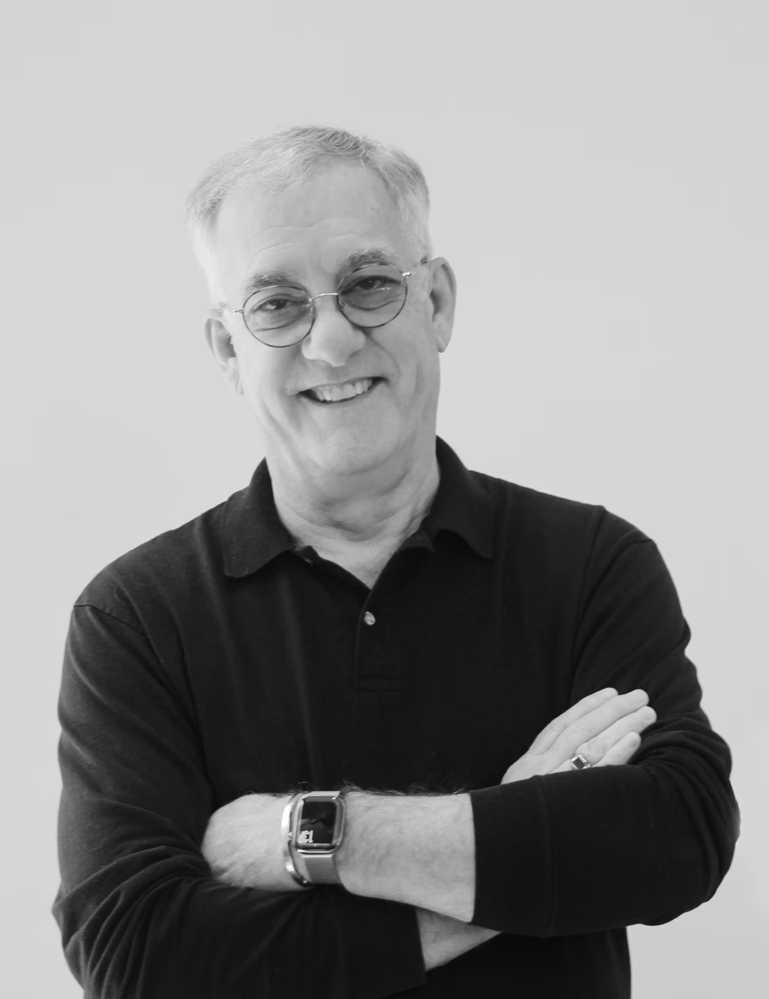 Smiling middle-aged man with glasses wearing a black turtleneck and watch, standing with arms crossed against a gray background.