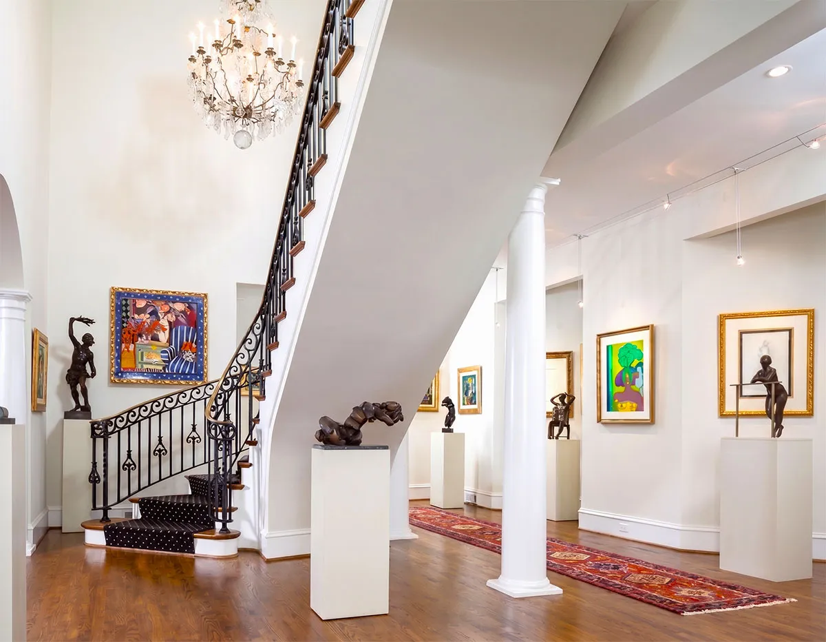 Bright art gallery with a chandelier, a black wrought-iron staircase, wooden floor, white walls with colorful framed paintings, multiple bronze sculptures on white pedestals, and a red patterned rug along the floor.