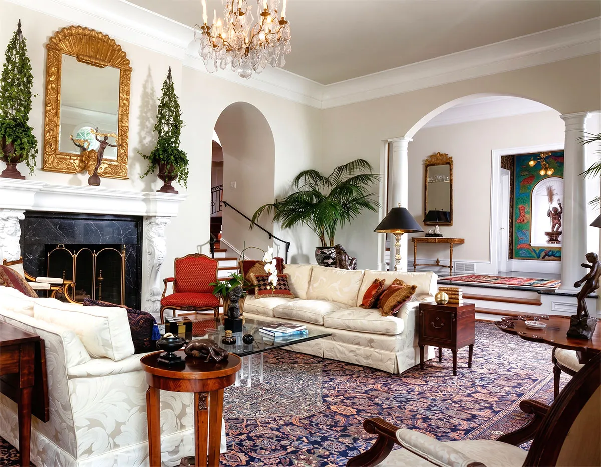 Elegant living room with cream sofas adorned with cushions, a glass coffee table on a patterned area rug, red upholstered chairs, a white ornate fireplace with a gold-framed mirror above, tall green plants, classic wood side tables, and a crystal chandelier hanging from the ceiling.